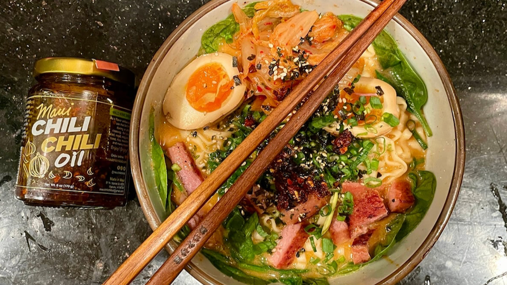 Bowl of ramen with chopsticks and a jar of chili oil on a dark surface