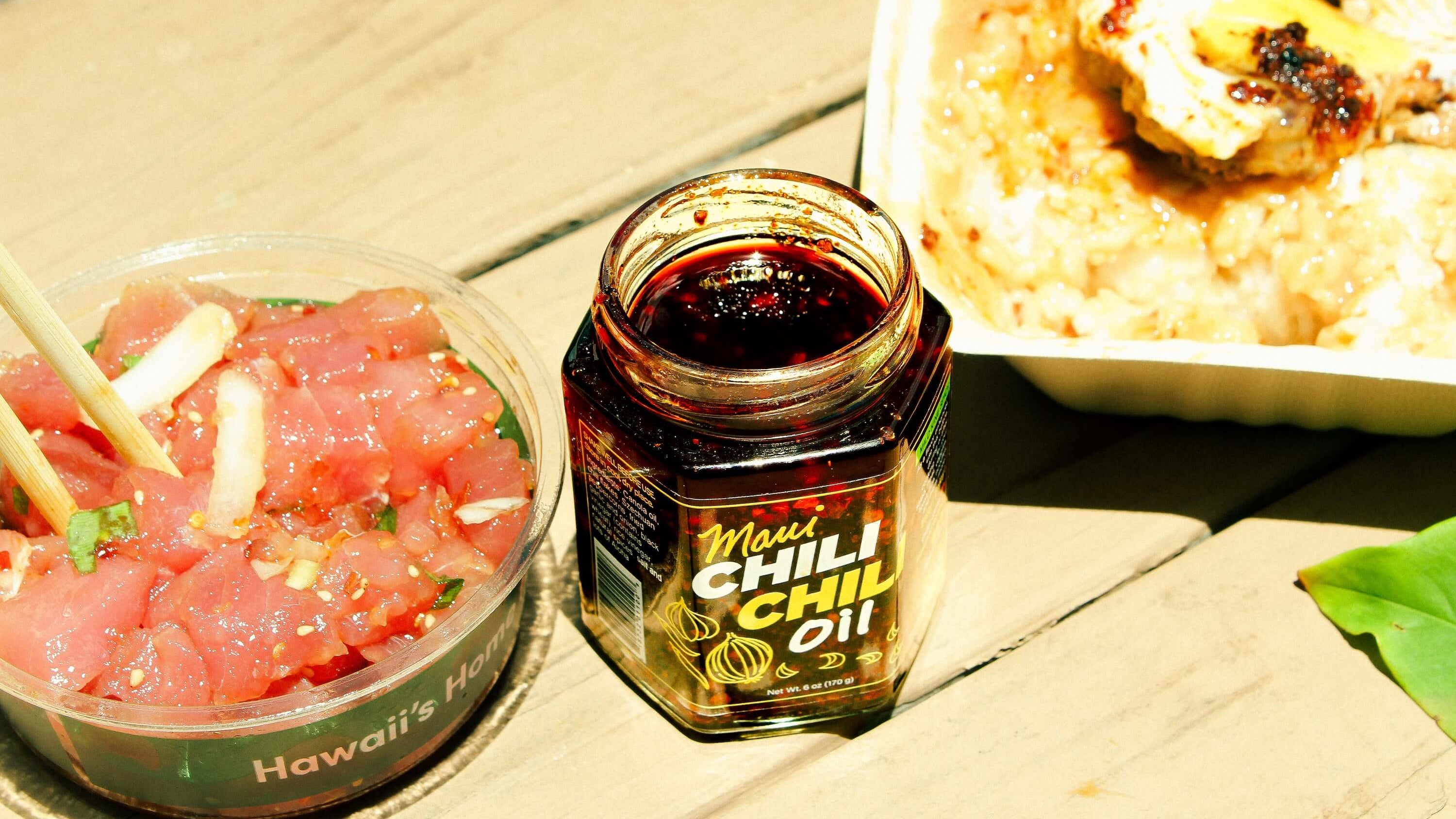 Jar of chili oil next to a container of raw poke and rice on a wooden surface