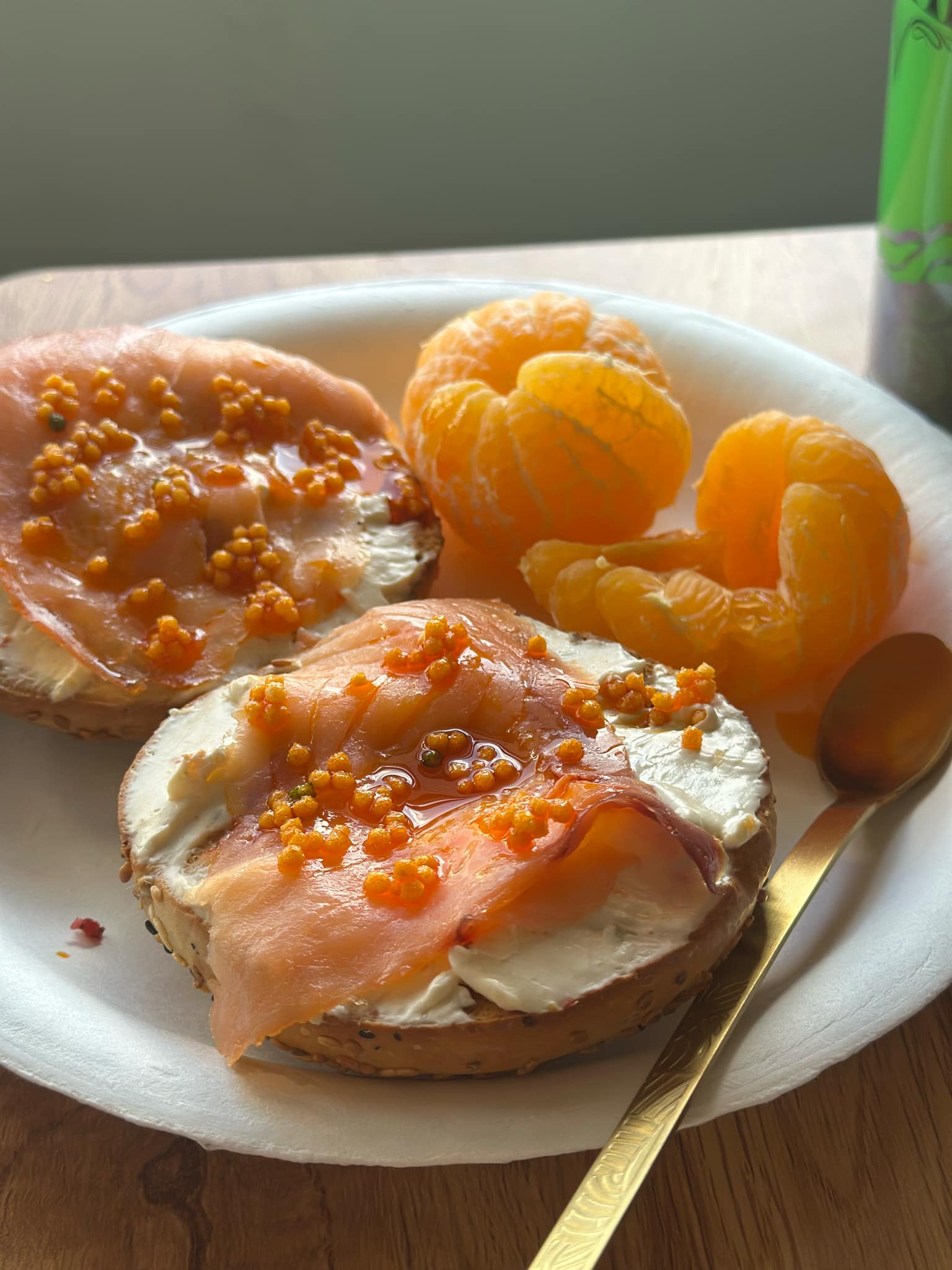 Bagel with salmon and cream cheese with bubu arare on a plate with mandarin oranges and a spoon.