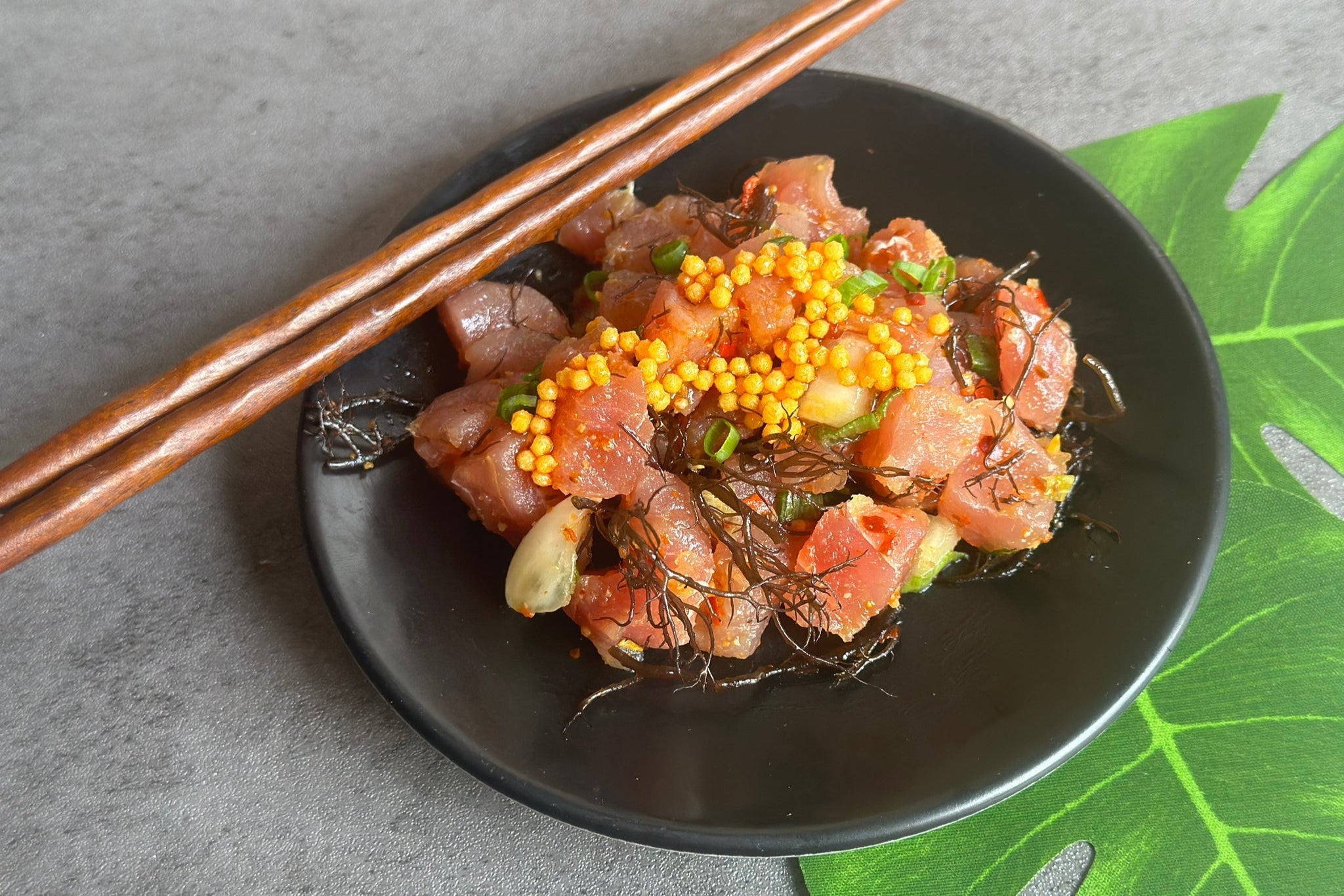 Black plate with raw fish dish garnished with yellow bubu arare and green herbs, accompanied by wooden chopsticks on a gray surface.