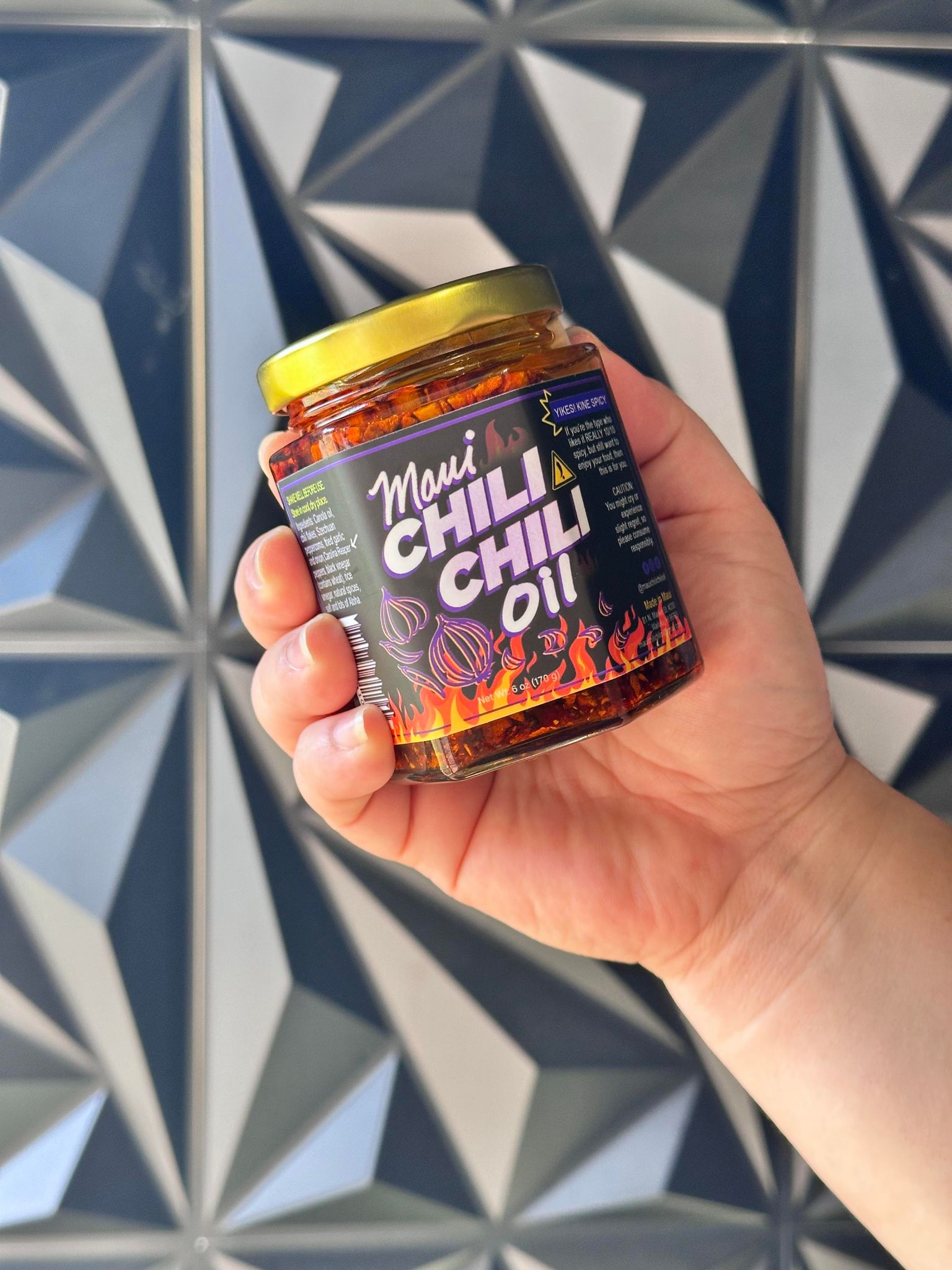Hand holding a jar of Maui Chili Chilli oil against a geometric-patterned wall.